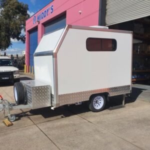 A white dog grooming trailer with a window and metallic accents is parked on a concrete surface outside a building with a pink wall and blue roller door labeled "Middy's." An open metal structure and a parked white vehicle are in the background.