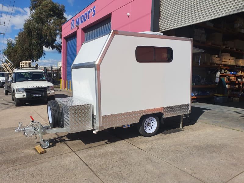 A white dog grooming trailer with a window and metallic accents is parked on a concrete surface outside a building with a pink wall and blue roller door labeled "Middy's." An open metal structure and a parked white vehicle are in the background.