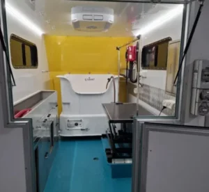 Interior of a mobile veterinary clinic with a bright yellow and white color scheme. Features include a stainless steel examination table, several cabinets for storage, and a mounted fire extinguisher. The floor is blue.