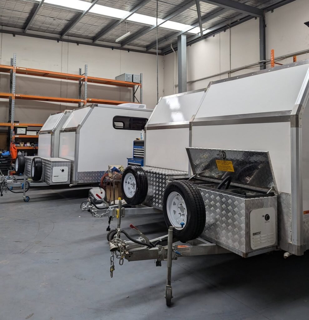 Two white trailers are parked inside a warehouse. They have diamond-patterned metal compartments at the front, each with a spare tire. Shelving and tools are visible in the background, indicating an industrial setting.