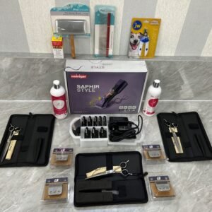 The Premium Dog Grooming Starter Bundle displayed on a table includes the Heininger Saphir Style clipper, scissors, combs, blades, cleaning solutions, and colorful dog supplies packaging. Items are neatly arranged in a well-lit space with a patterned wall backdrop.