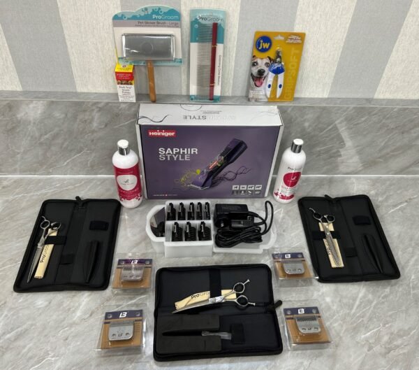 The Premium Dog Grooming Starter Bundle displayed on a table includes the Heininger Saphir Style clipper, scissors, combs, blades, cleaning solutions, and colorful dog supplies packaging. Items are neatly arranged in a well-lit space with a patterned wall backdrop.