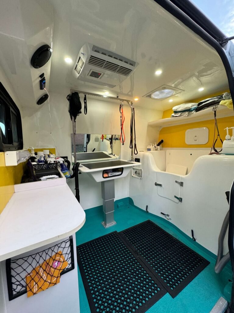 The image shows the interior of a brightly lit grooming van. It features a grooming table, sink, storage shelves, and a turquoise floor with black non-slip mats. The walls are white with yellow accents, and grooming tools are visible.