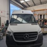 K9 Trailer Rentals A white Mercedes-Benz van is parked inside a garage with a high ceiling and shelving units. The garage has a windowed roof that lets in natural light. The van's front view, including its grille and headlights, is visible.