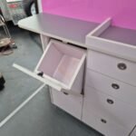 K9 Trailer Rentals A medical or veterinary cabinet with a stainless steel surface and a pink back panel. One open compartment reveals a pull-out white trash bin. The cabinet has multiple closed drawers with round metal handles.