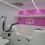 K9 Trailer Rentals The image shows the interior of a brightly lit, modern pet grooming station. It features a deep tub, grooming table, adjustable shower head, and a prominent pink accent wall. Overhead lights and a ventilation fan enhance the clean, functional space.