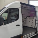 K9 Trailer Rentals White van with a sliding side door open, revealing an empty interior with built-in shelving and cabinetry. The vehicle is inside a garage with a partially visible roll-up door and gray concrete floor.