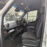 K9 Trailer Rentals Interior view of a white van's cabin, showing two black seats, a steering wheel, and a dashboard with various controls. The driver's side door is open, and the floor is covered with a dark mat. Background shows a workshop setting.