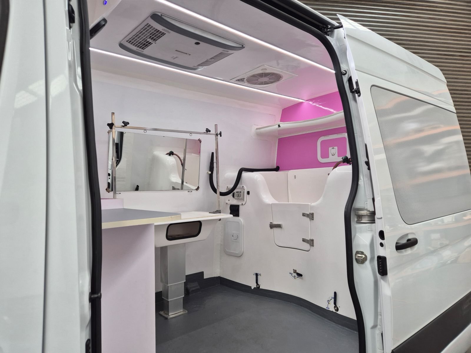 K9 Trailer Rentals The interior of a white van converted into a mobile clinic or workspace, featuring a counter, mirror, hooks, shelves, and pink accent paneling, with the side door open and bright lighting overhead.