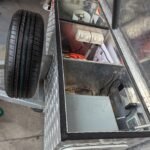 K9 Trailer Rentals A metal utility box with an open lid shows electrical outlets, wiring, and a control panel inside. A spare tire is mounted on the side, and workshop tools and equipment are visible in the background.