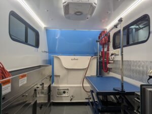 K9 Trailer Rentals Interior of a grooming trailer features stainless steel cabinets, a blue examination table, medical equipment, overhead lighting, and warning labels—perfect for those seeking a grooming trailer for sale Melbourne or considering trailer rentals.
