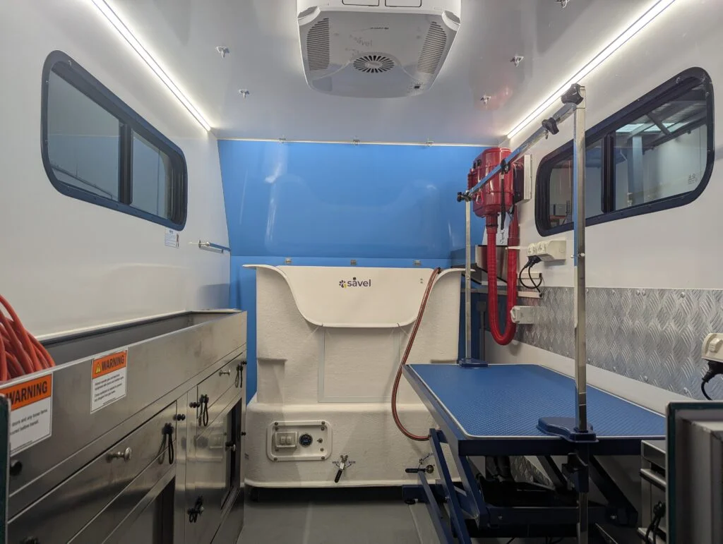 K9 Trailer Rentals Interior of a grooming trailer features stainless steel cabinets, a blue examination table, medical equipment, overhead lighting, and warning labels—perfect for those seeking a grooming trailer for sale Melbourne or considering trailer rentals.